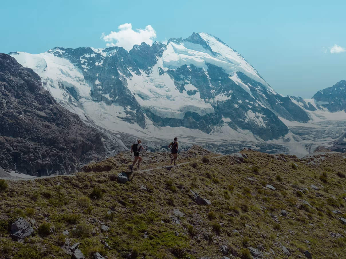 Two people running through a Swiss mountain landscape, with snow-covered peaks. 
